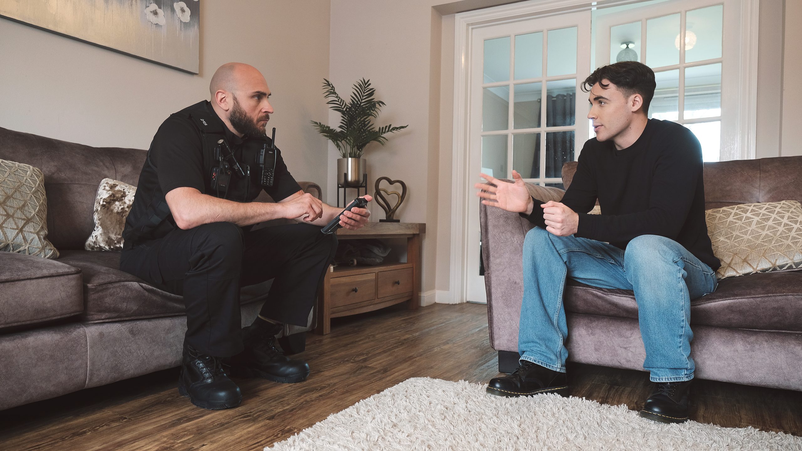 two people sit in a sitting room having a conversation. The person on the left is wearing a police officer uniform, and the one on the right is wearing a black t-shirt