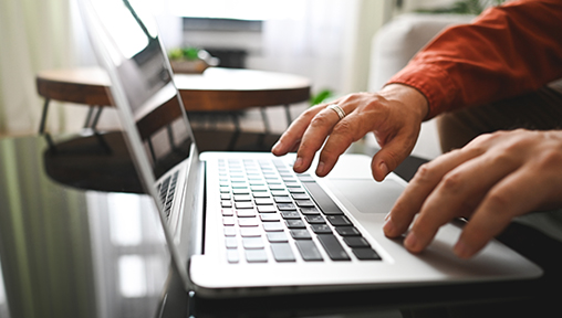 an image of a silver laptop, with two hands to the right of the image reaching toward the keyboard to type. the person is wearing an orange shirt.